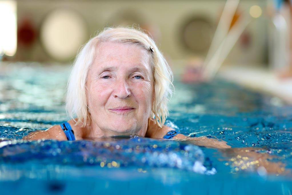 Image: a day in the life of a blind person. An older person with shoulder length hair is swimming in a pool next to the side of the pool.