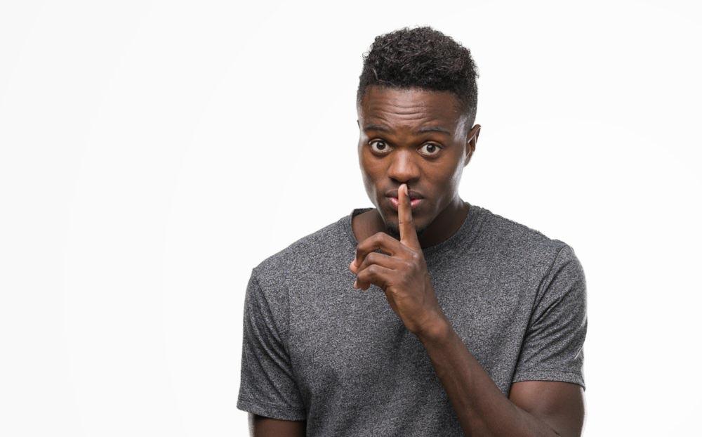 Photo for the article: What to say when someone says they have a disability? Young Black man wearing grey t-shirt asking to be quiet with finger on lips. 
