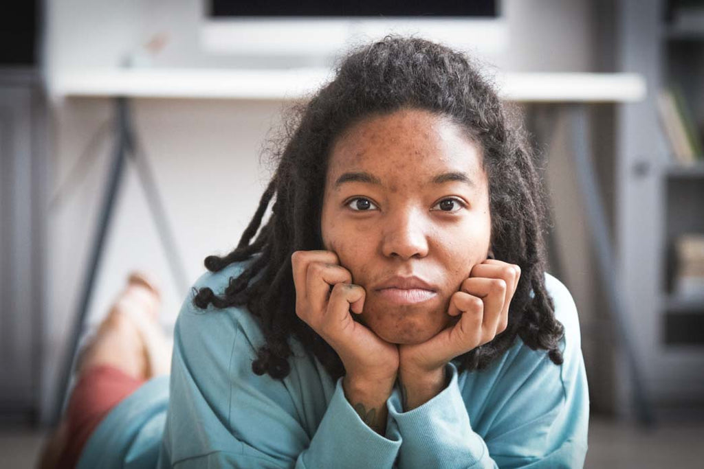 Black women and anxiety: a color photo portrait of a Black woman with anxiety looking directly at the camera. She is lying on the floor in a modern lounge room with her head resting on her hands. She is not weaing make up.