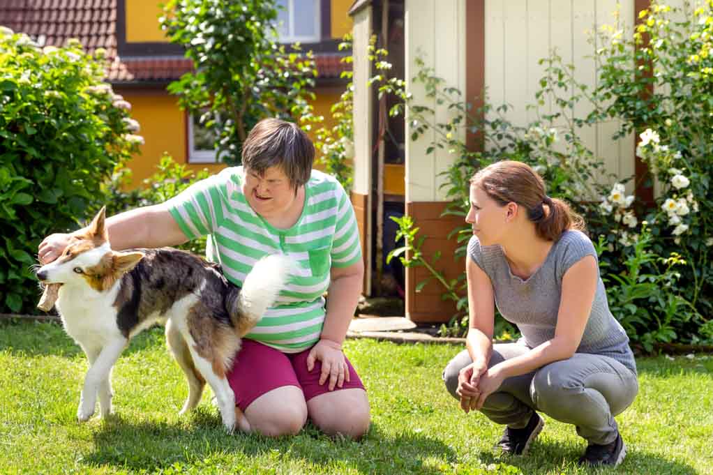 What is the correct term for disabled person? Two people are having a conversation on a green lawn, while a pet dog plays with them. One of the people is disabled.
