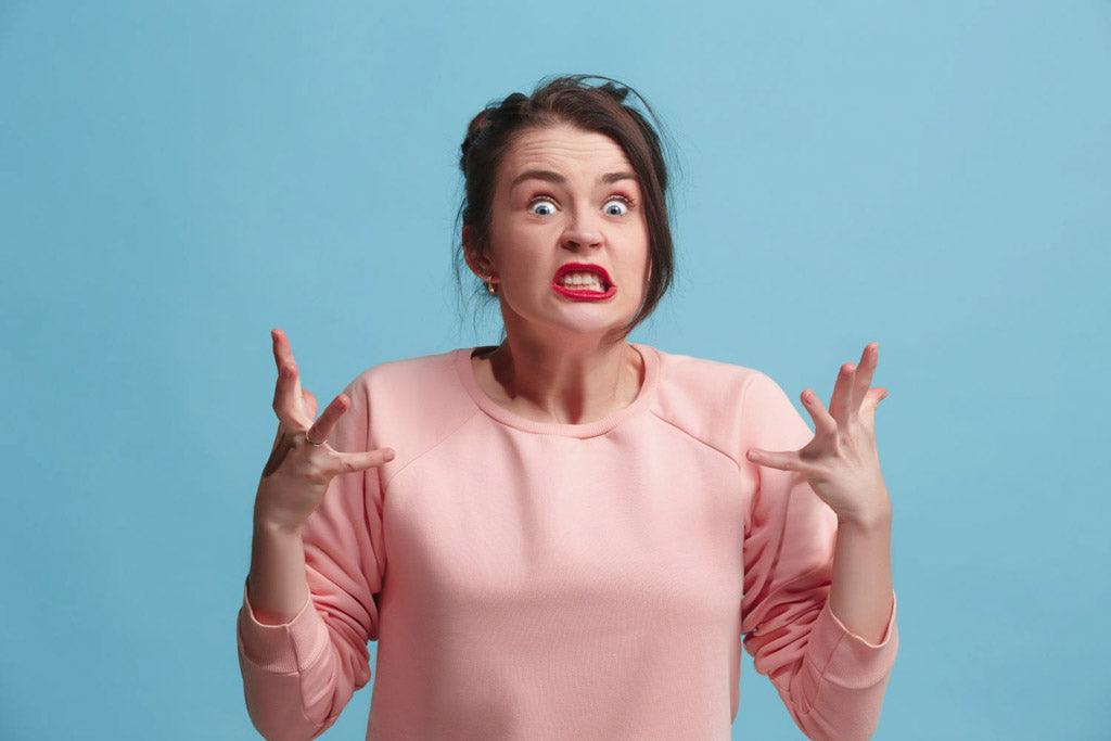 Person in a pink sweater with raised hands and a grimacing expression against a blue background.