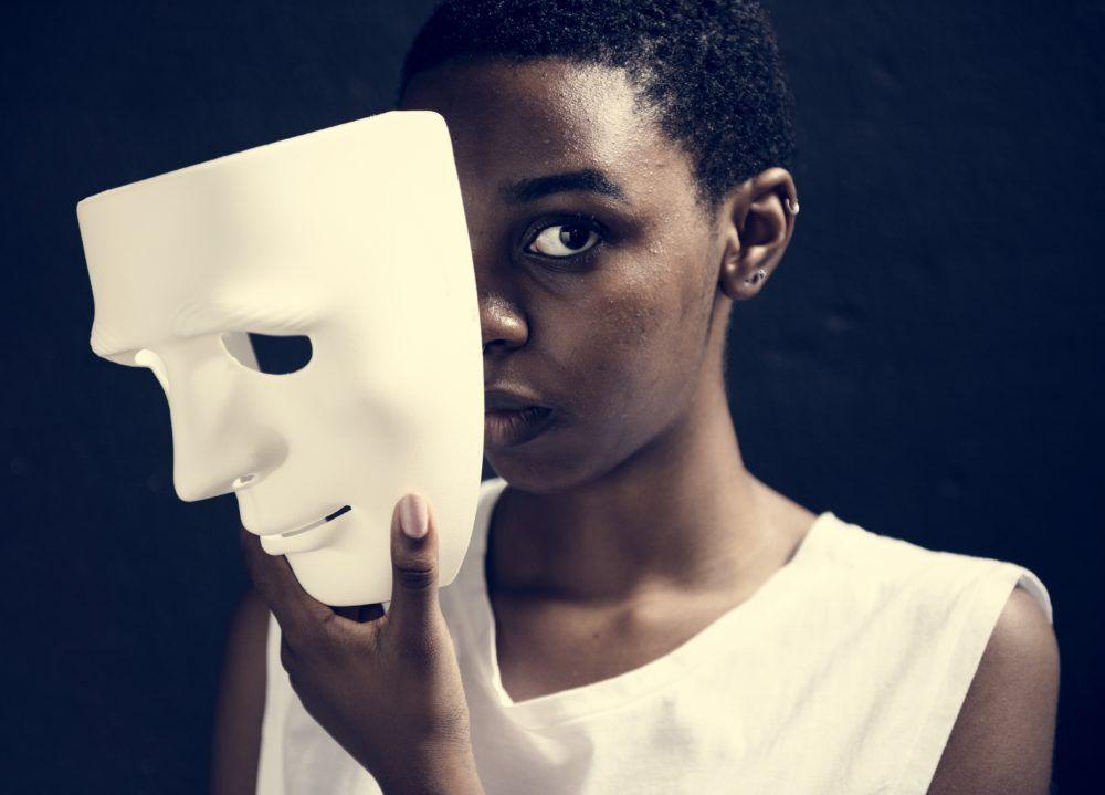 Young person holds a white mask to their face so that it covers less than half. They have piercing eyes that look straight at the camera. For article on what not to say to someone with bipolar disorder.