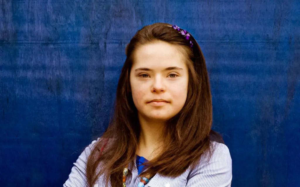 Image for an article about bias against people with Down syndrome. Portrait of a teenage girl with Down's syndrome standing against a blue wall.
