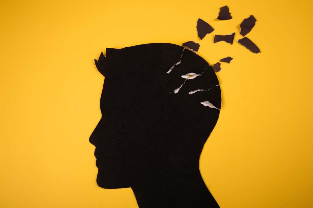 A paper cutout illustrating what does a coma feel like: a silhouette of a person's head is set against a yellow background; pieces of their head are detached and floating outside the body.
