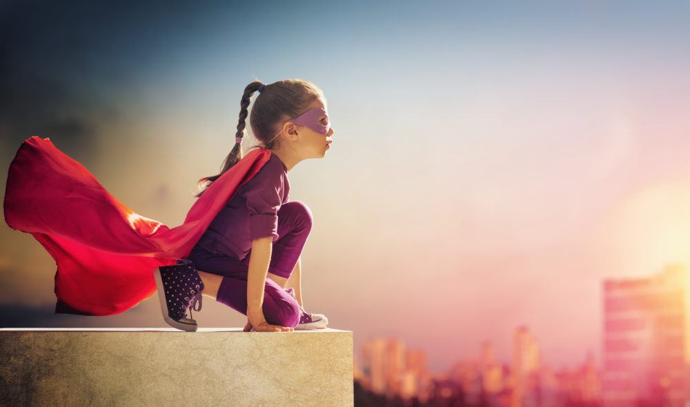 ADHD is my superpower. Little girl wearing a purple tracksuit and pink cape and mask, looks out across the horizon perched on a rooftop. She is crouched down, has a look of super focus, looking as if she is about to jump off and fly. 