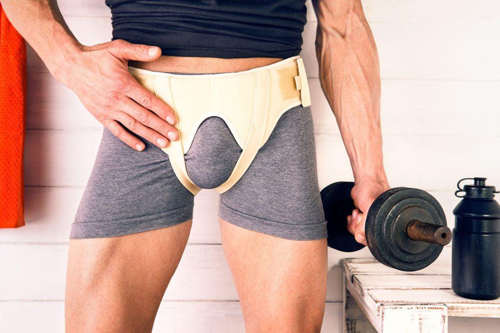 Front photo of a male torso from the middle of the chest to the knees. He is holding a dumb belll in his left hand and is wearing an inguinal hernia bandage. Image for an article answering the question: 