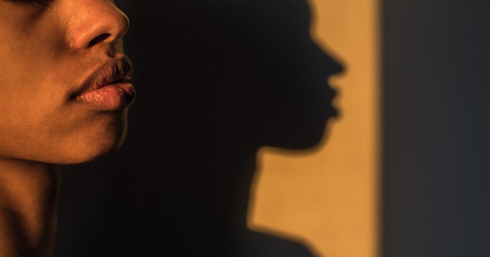 A photo of a person's face, with only the nose, lips and chin visible. Their face casts a shadow a wall. The photo is show at sunset with yellow hues. Photo for an article "Why I became a sex worker."
