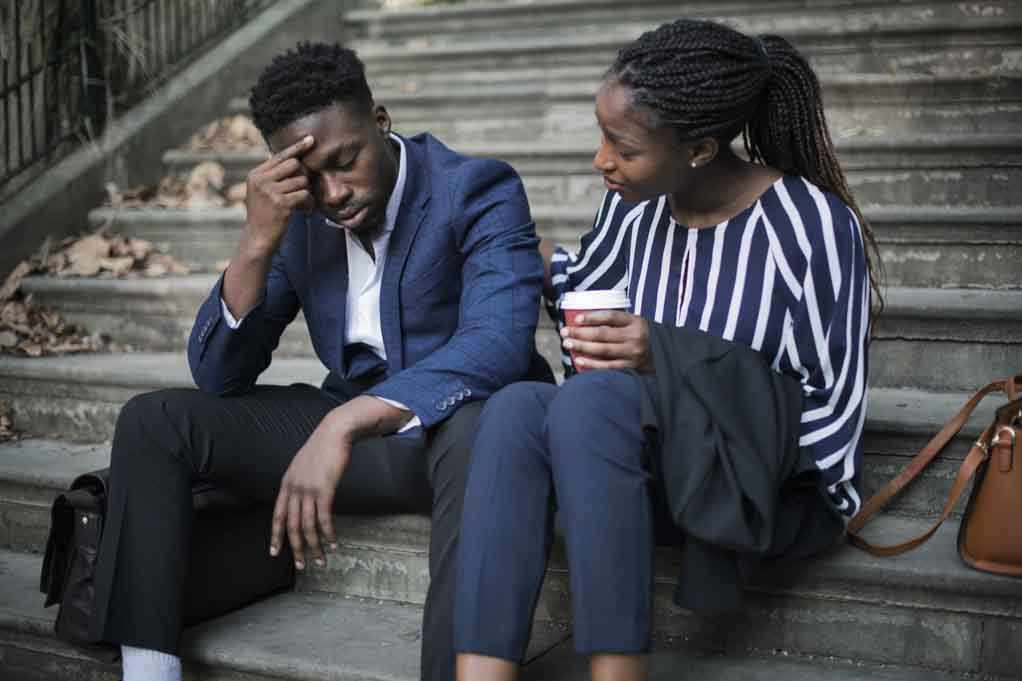 Showing how to support someone, an African-American woman listens and comforts a male friend. They are sitting side by side.