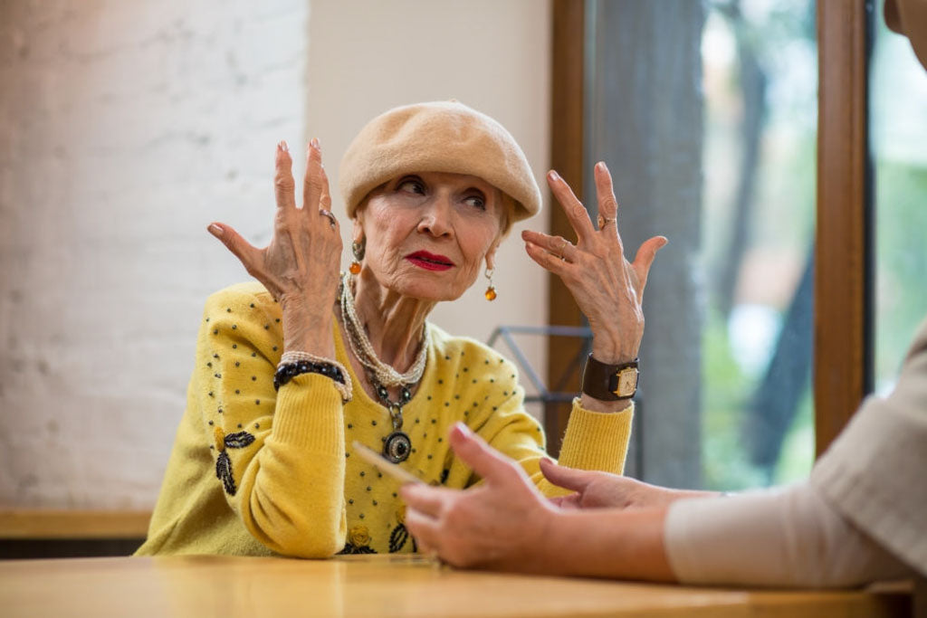 Picture of Grandma Stella dressed in yellow, with a cap on her hand, gesticulating with her hands in response to personal questions about breast cancer.