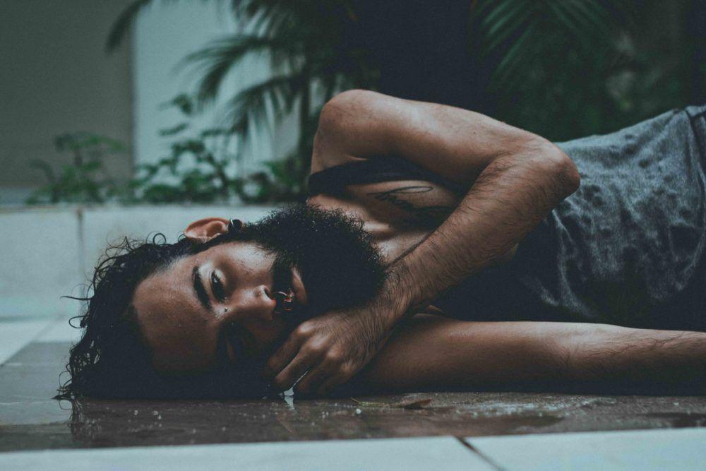 A person is lying on their side looking at the camera. They have a beard and are wearing blue bib overalls. Their face looks sad. 