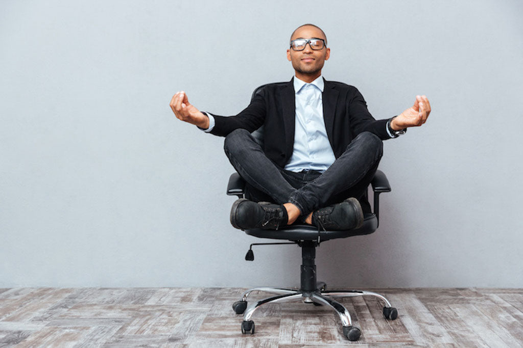 Returning to work after cancer: why is it so hard? Relaxed young man sitting and meditating on office chair. He wears black-rimmed glasses, a black blazer