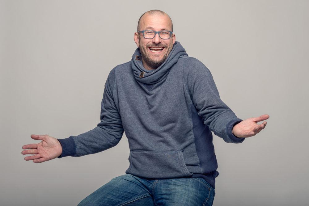 A seated person wearing blue glasses and a grey hoodie laughs with both hands held open.