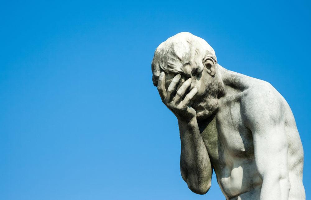 Stone sculpture of a man covering his face with one hand against a clear blue sky.