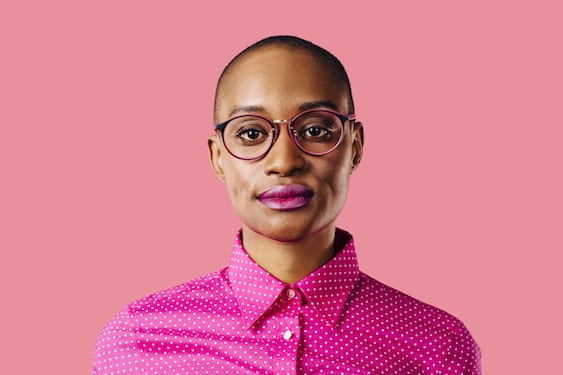 Photo for article on should I tell my clients I have cancer, Photo of androgynous woman with buzzed cut and large rimmed glasses in pink shirt against pink background.