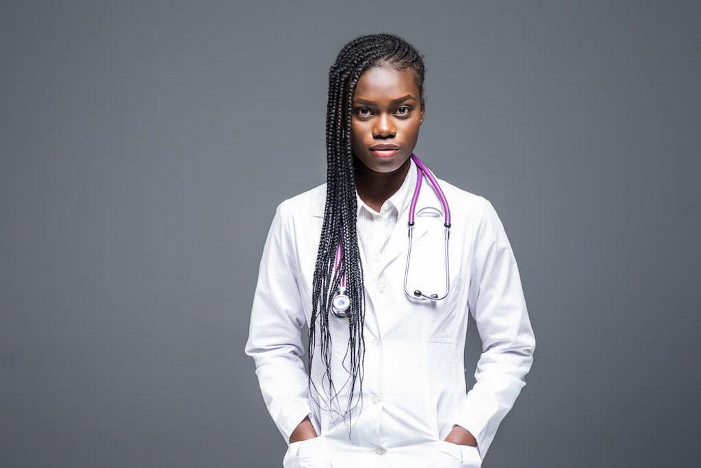 Photo for an article on why some people of color don't trust doctors. A doctor with long corn-row braids in a white coat with a pink stethoscope looks intently at the camera.