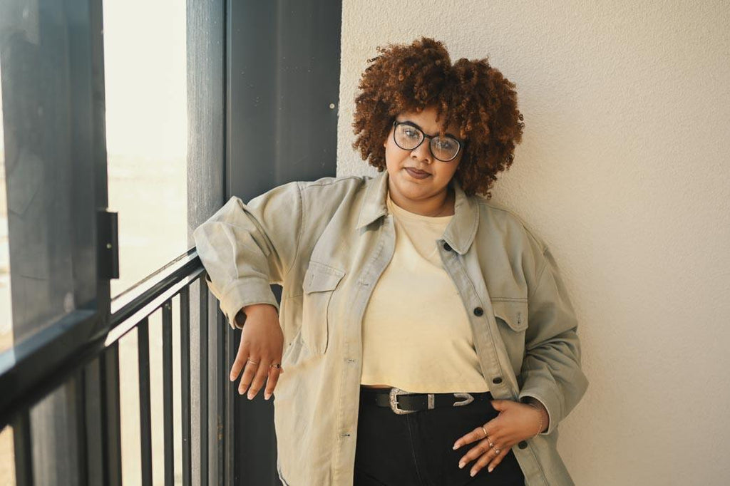 What does it mean to choose our own bodies? A color photo of a happy smiling curvy black woman with afro hair posing in beige t-shirt, jeans, stylish glasses on a sunny balcony.
