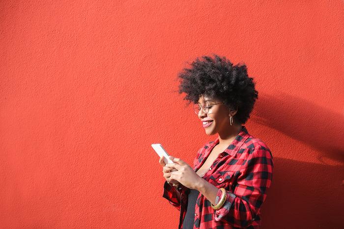 Image for article: 'What are the benefits of wishing someone well?' Young cheerful woman with an afro and glasses smiles at her phone