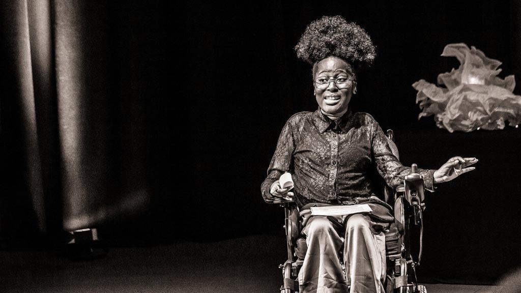 Black and white photograph with a warm tone. The image shows the disabled musician Miss Jacqui seated in a wheelchair against a black curtain and lit by a spotlight. She is performing to an audience. She has been captured 'in action.