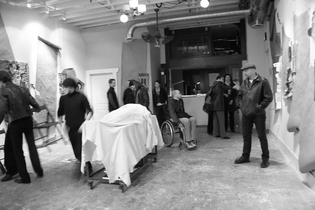 A black and white photo of people walking around a room. In the middle of the room is the body of a person, on a hospital gurney, draped in a white sheet.