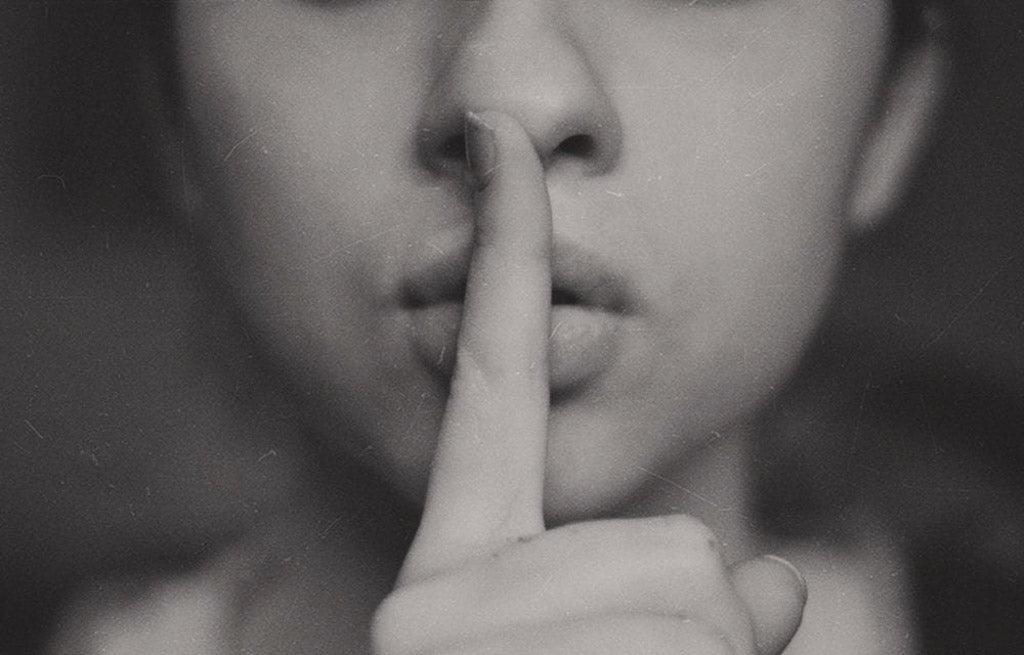 Photo for article: Talking to doctors about the side effects of psych meds. Sepia close up photo of a woman's face. She is placing her finger over her mouth indicating 'be quiet'