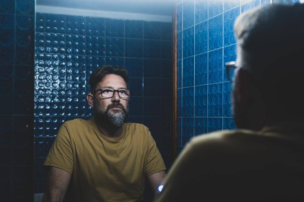 Nightime panic attack: a photo of a bearded person with glasses staring at a bathroom mirror at night. They look anxious and depressed.