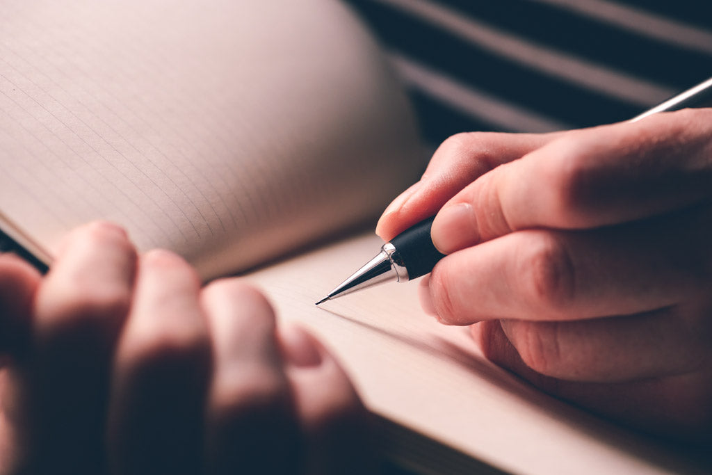 Image for an article about learning to write left handed. Photo of a woman in her writing diary with her left hand, close up of hands with pencil and notebook.
