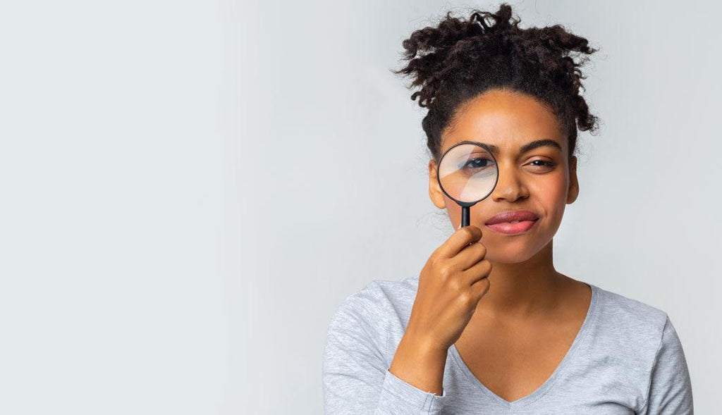 Myths about celiac disease. A photo of a curious Black woman holding a magnifying glass over her right eye while looking directly at the camera.