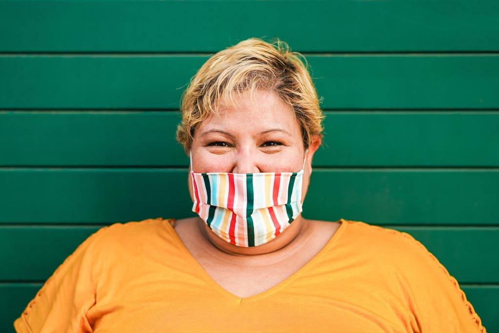 Why I hide my chronic pain? A person is standing against a green wall wearing a colorful mask to hide their pain.