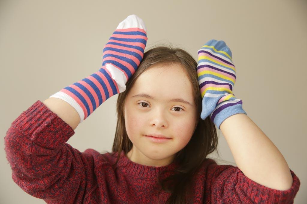 A picture of a daughter diagnosed with down syndrome smiling at the camera. She is holding her hands up against the side of her head like rabbit ears. On each hand she has a different coloured sock.