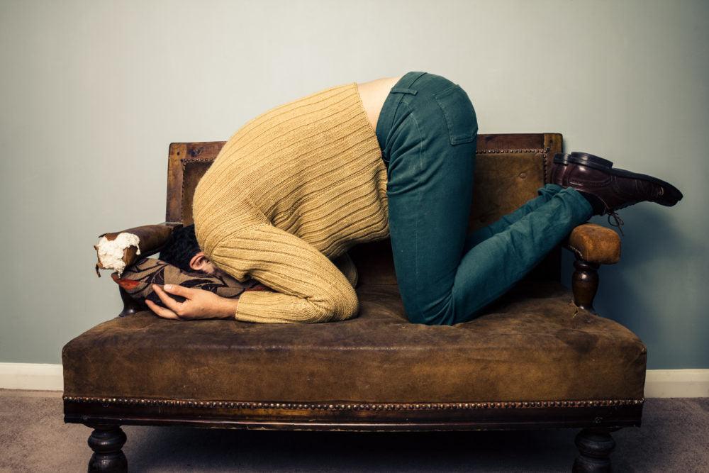 Dying from alcoholism: what is it like? Photo of a person burying their face into a brown large armchair. The person wears a yellow sweater, blue jeans and brown leather shoes. 