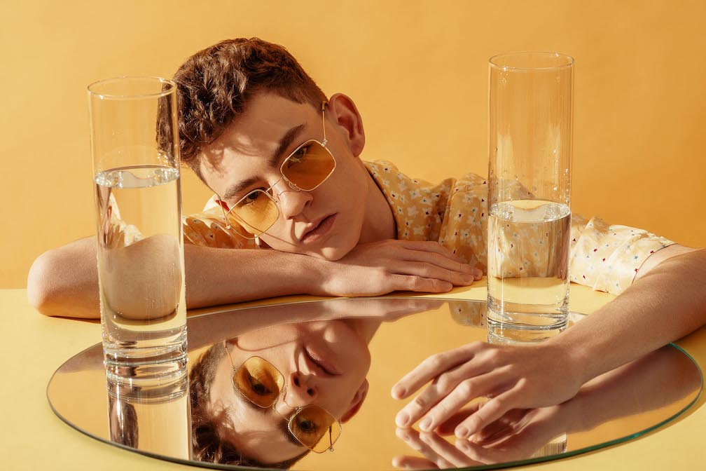 Is self diagnosis valid? A photo of a fashionable young man wearing trendy sunglasses, posing on yellow background. Their face is reflected into a mirror.