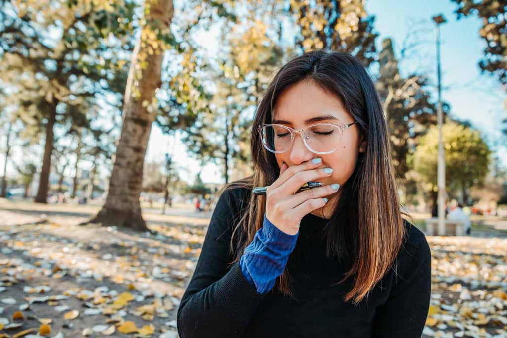 Living with ulcerative colitis: a person with long hair and glasses is sitting down in a park, eyes closed, their hand covering their mouth. There is a pen in the hand.