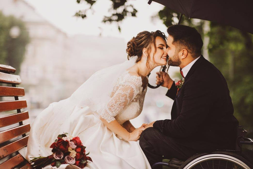 Love and disability. A bride in a white wedding dress, and the groom in a wheelchair, sit kissing on the bench in the park