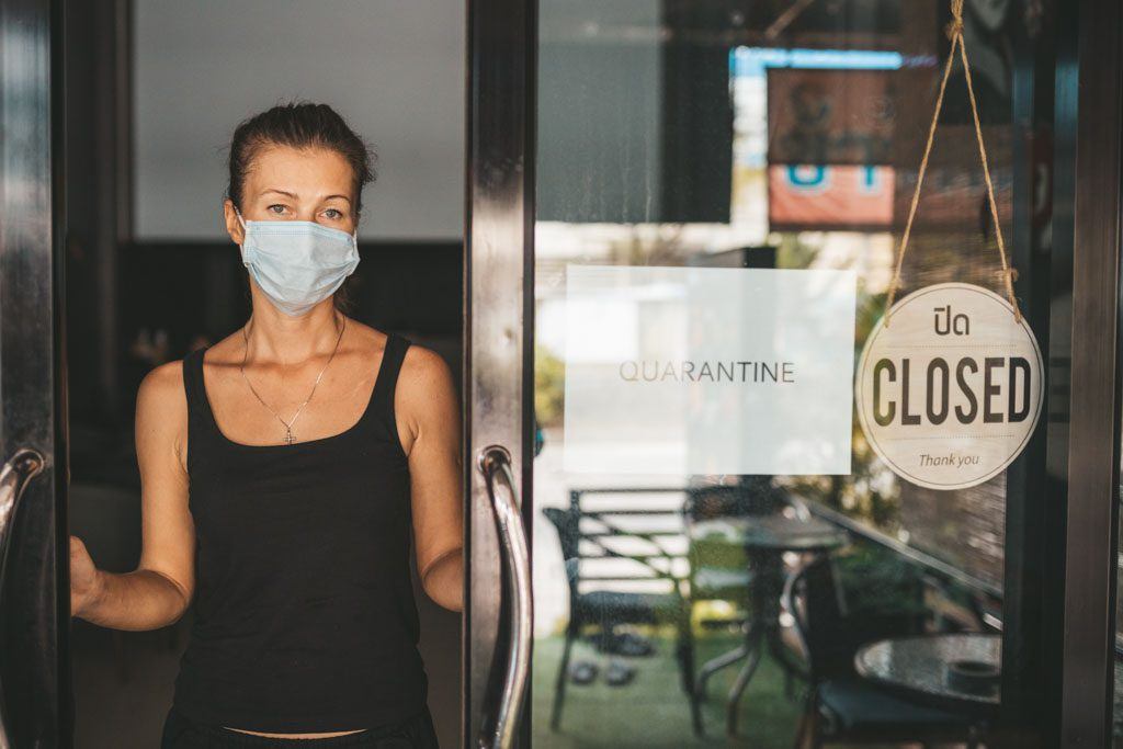 A woman restaurant owner, in medical mask, stands in the doorway of their closed restaurant for quarantine. Image for an article on missing dining out at restaurants because of the pandemic.