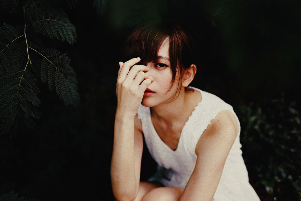 Image for article: Is self worth based on how someone looks?. Thin young Vietnamese lady looks pensively at the camera. Her right hand partially covers her face. She is crouching down under a tree.