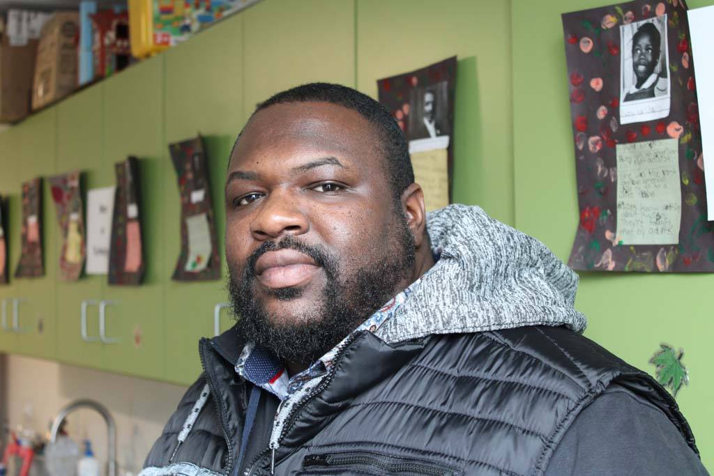 A photo of Darnell Hill sitting down looking at the camera with a serious expression. He is wearing a black winter jacket. Behind him are pictures of black teenagers affected by police violence.