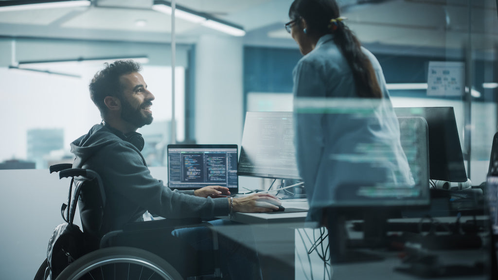 Seeking employment with a disability in tech: a disabled person in a wheelchair is sitting in front of computer screens talking to a colleague.