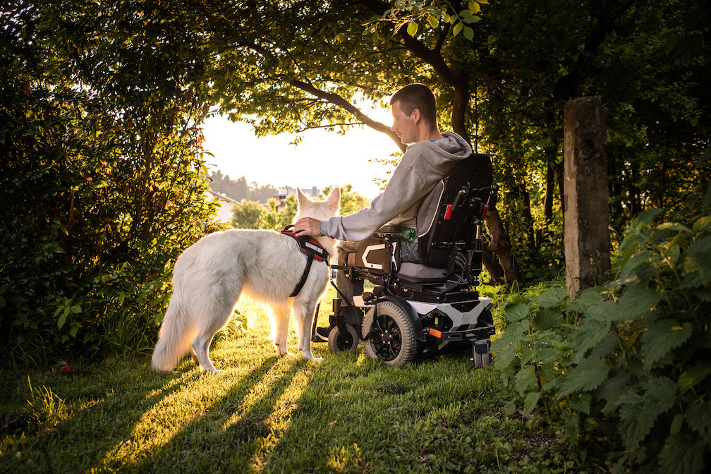 A man in a power wheelchair rests a hand on a white dog beside a garden gate.