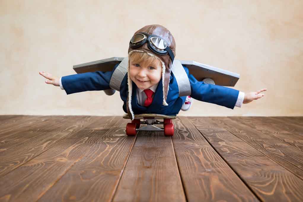 A child wearing an aviator cap and toy wings lies on a skateboard.