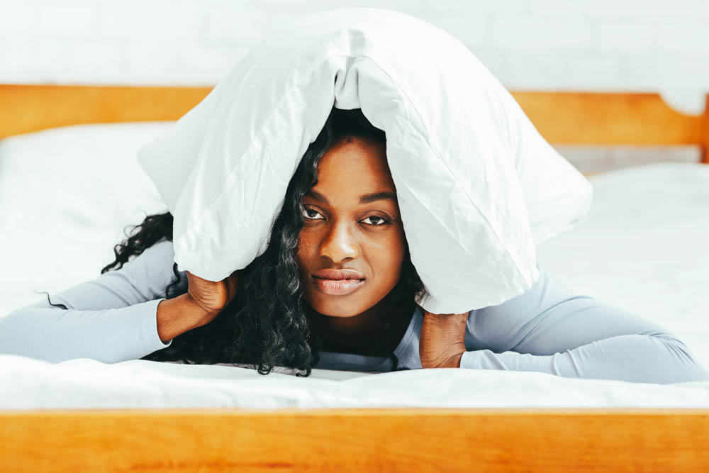 Article about the term 'crazy woman' - photo of a Black woman lying in bed on her stomach looking at the camera. She has a pillow covering her ears.