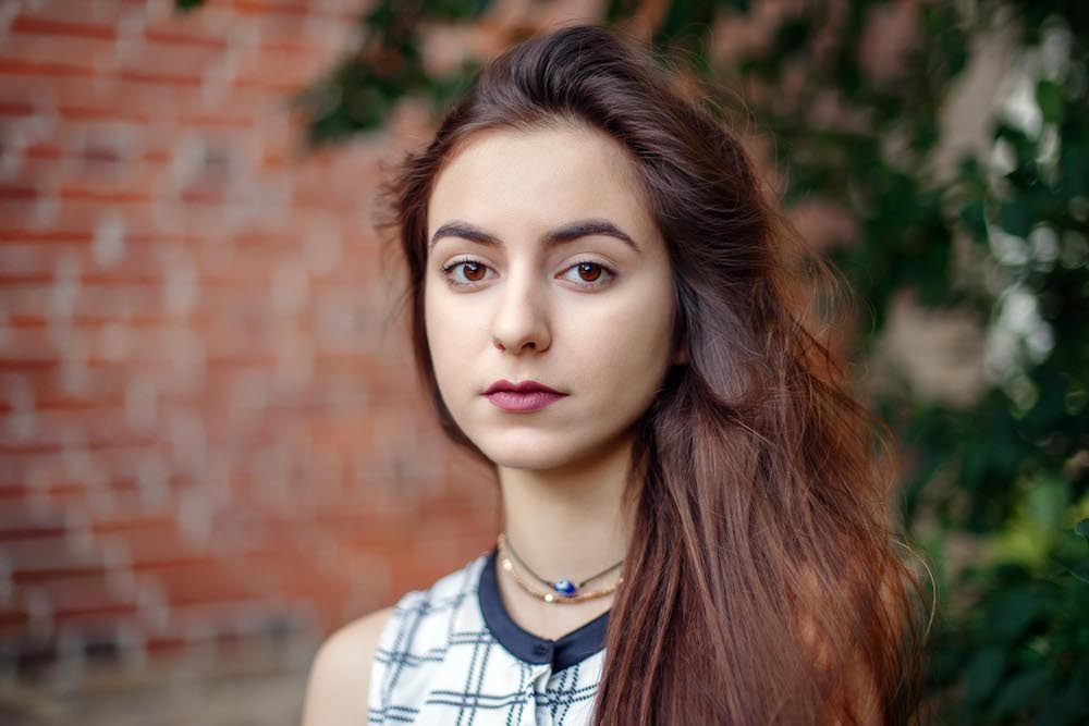 Stories about connective tissue disease: Closeup portrait photo of a pensive young woman with long dark hair, black brown eyes, looking directly into the camera. She is wearing a white plaid shirt and is standing against a brick wall.