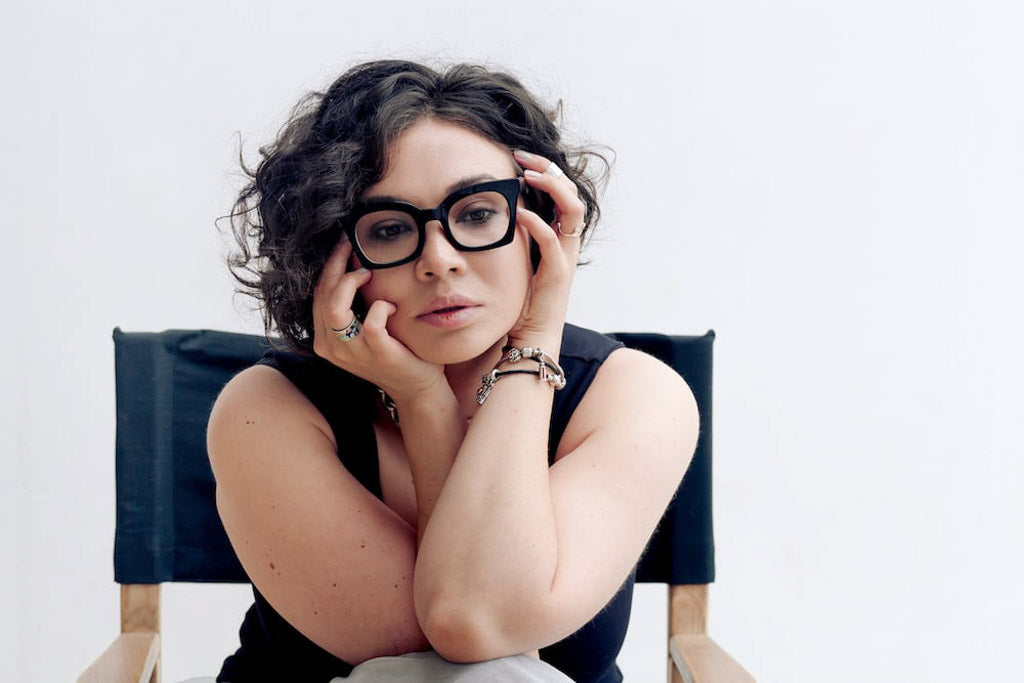 'Talking about dying with your doctor won't kill you.' Portrait of plus-size woman with dark curvy hair wearing fashionable glasses and bijouterie. Natural lighting, white background. She looks nervous and contemplative, eyes looking to the ground.