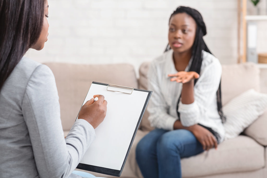 A person holding a clipboard sits opposite another person on a sofa.