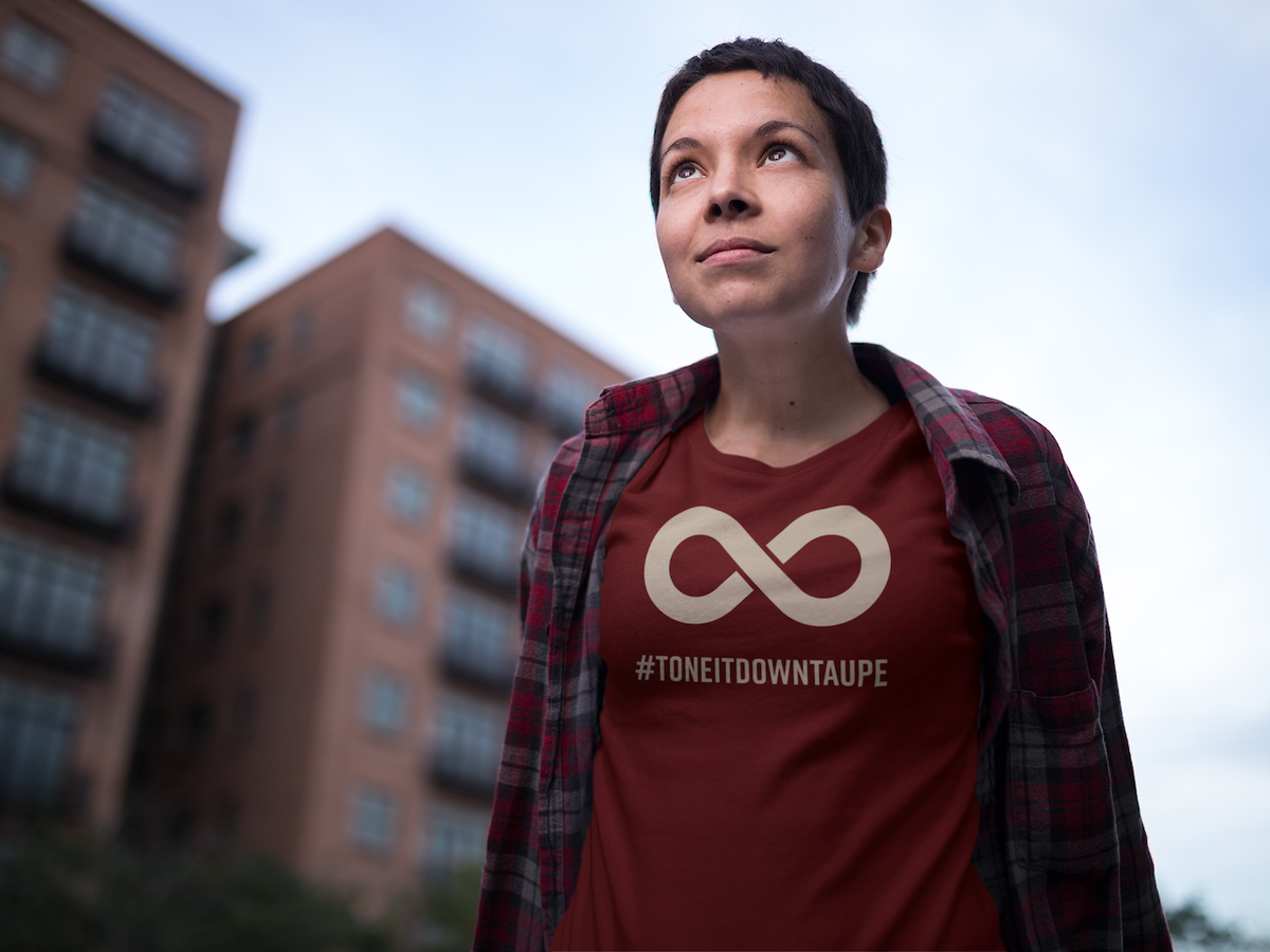Person outdoors wearing a burgundy Tone It Down Taupe T-shirt with a taupe infinity symbol and #ToneItDownTaupe text.