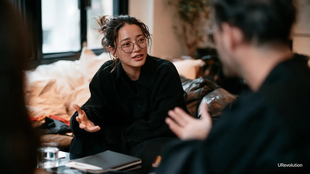An adult woman speaks with another person in a warm, quiet room, with a notebook and glass of water nearby.