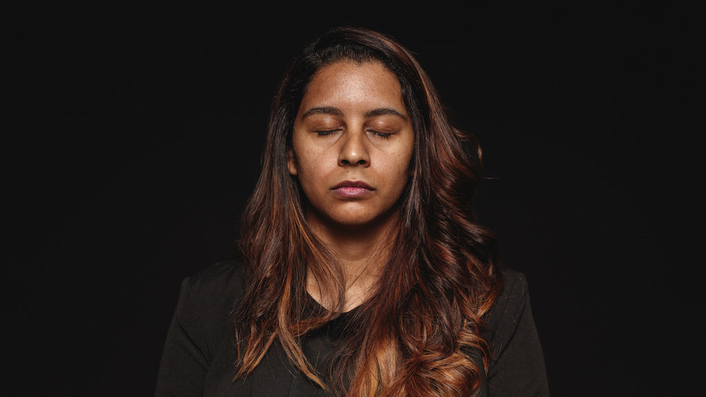 What does an emotional wound look like? A woman with long flowing brown hair is looking at the camera with their eyes closes. They are standing against a black background.