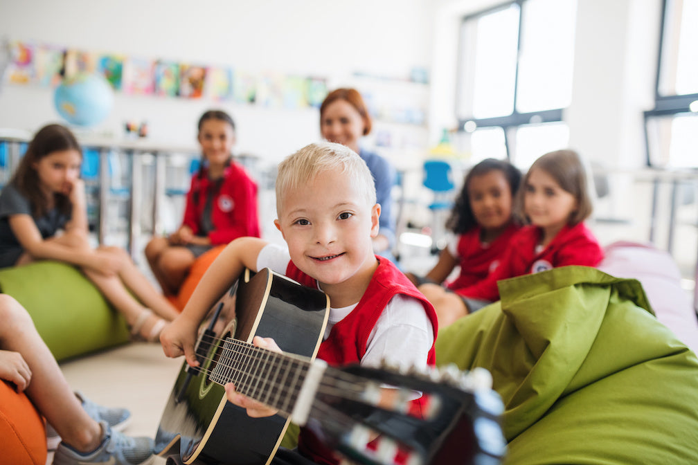 What does inclusive education mean? A photo of a down-syndrome boy with school kids and teacher sitting in class, playing guitar.