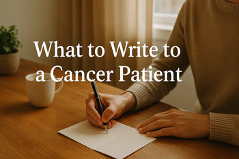 A person sitting quietly at a table, writing a card by hand. There’s a soft light coming from a nearby window, suggesting warmth and contemplation. On the table: an open card, a pen, a cup of tea or coffee, and perhaps a small plant or candle. No faces ar