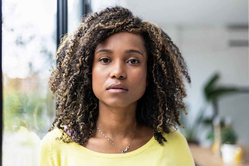 A woman with curly hair wearing a yellow top, seated beside a window and looking at the camera.