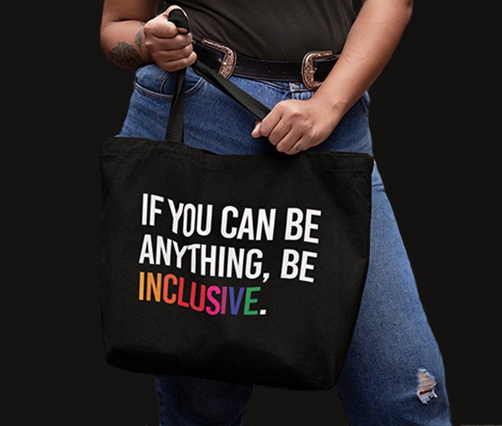 A close up of a person wearing jeans against a black background. They are holding a tote bag with the phrase, "If you can be anything, be inclusive," printed on the front of it.